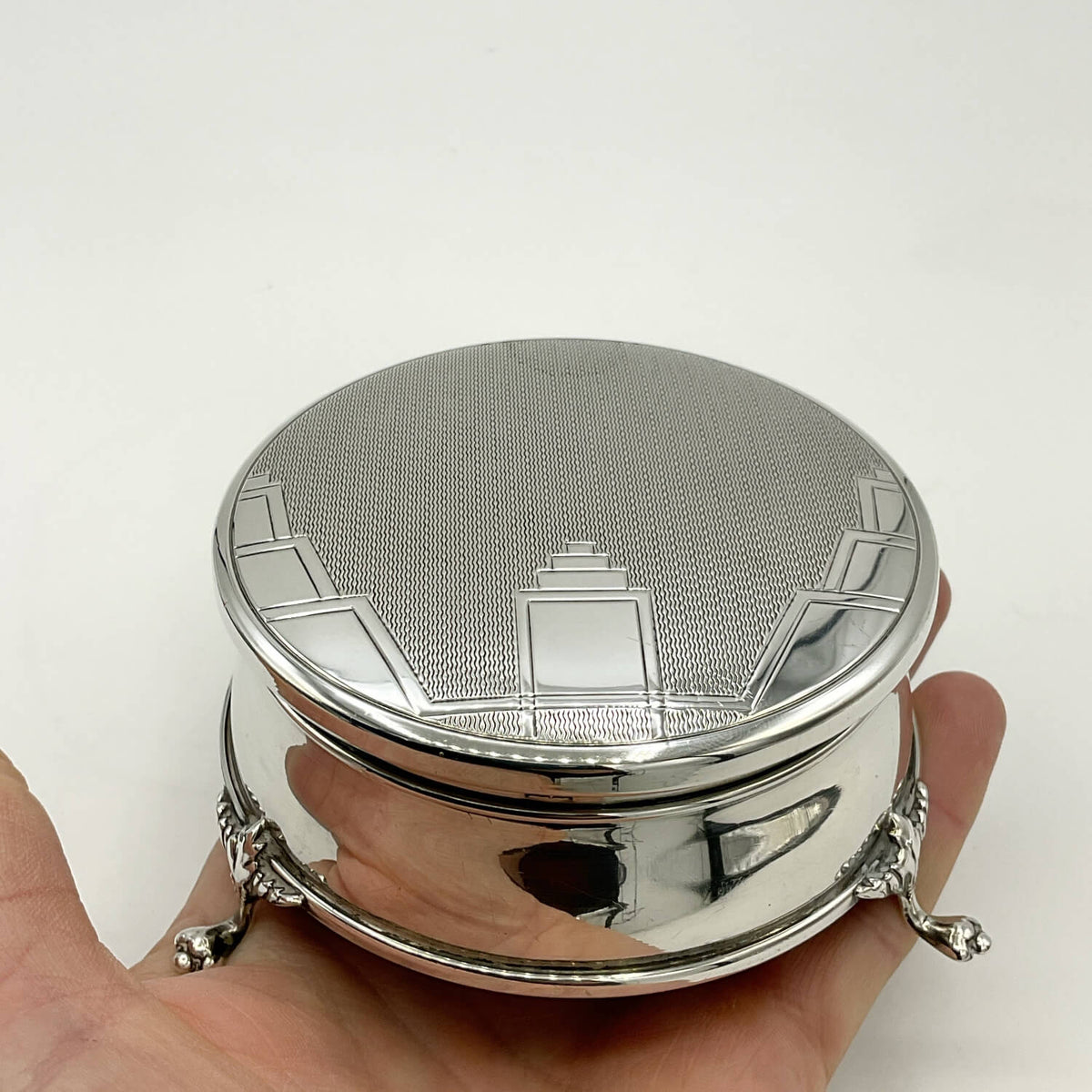 A polished silver trinket box with an Art Deco design lid and small decorative feet, shown resting on a person's hand against a plain light background.