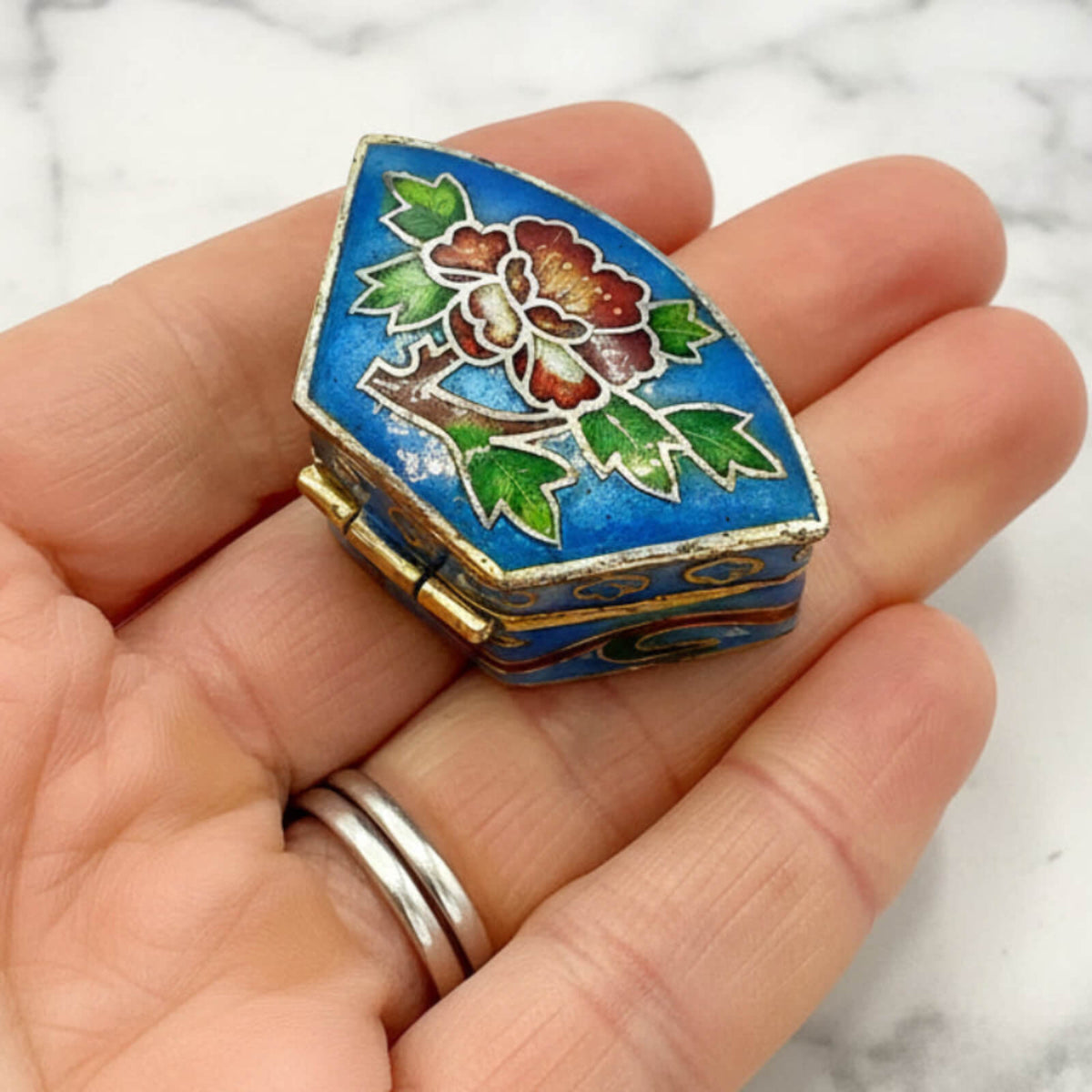 Small decorative box with floral design held in a hand against a marble background
