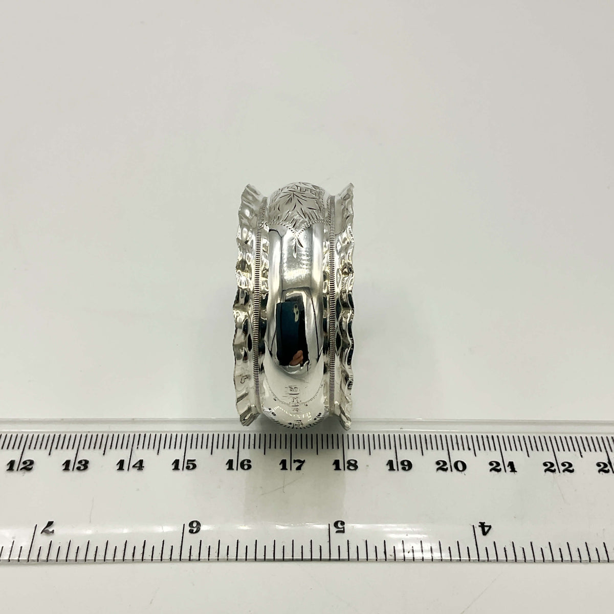 Silver napkin ring with intricate design on a white background with a ruler for scale.