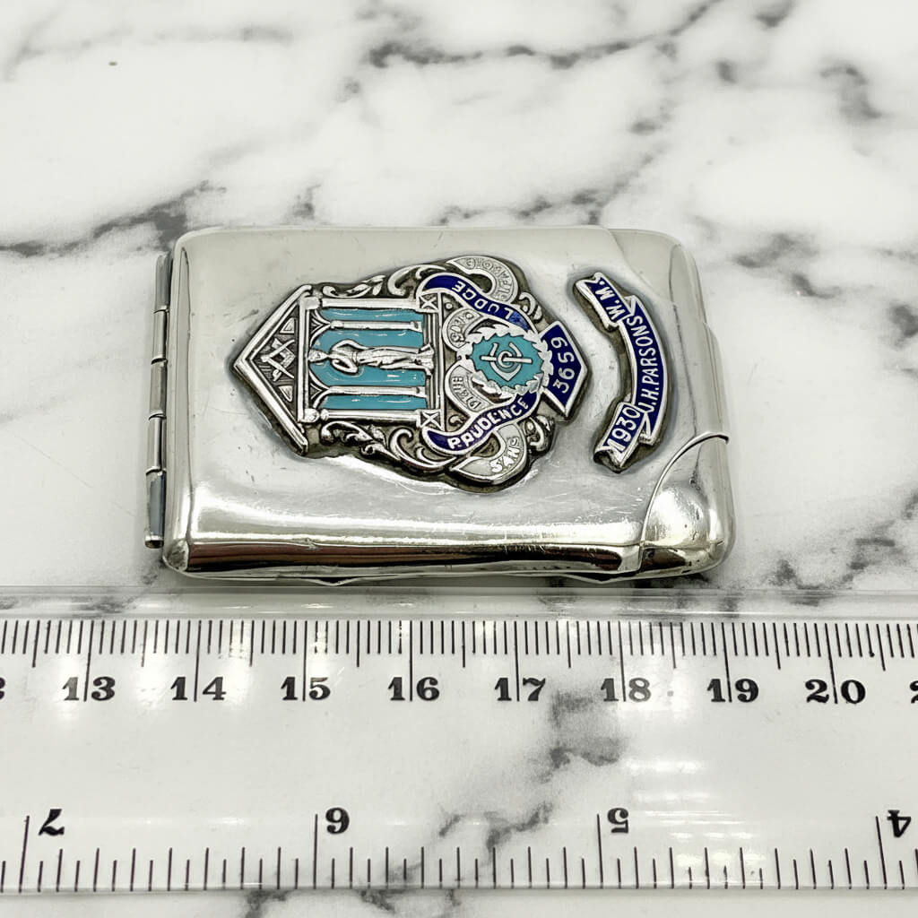 Silver matchbook with emblem and text on a white background, measured against a ruler.
