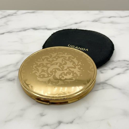 Gold powder compact with a design to the lid and a black bag behind with Uganda imprinted on it on marble