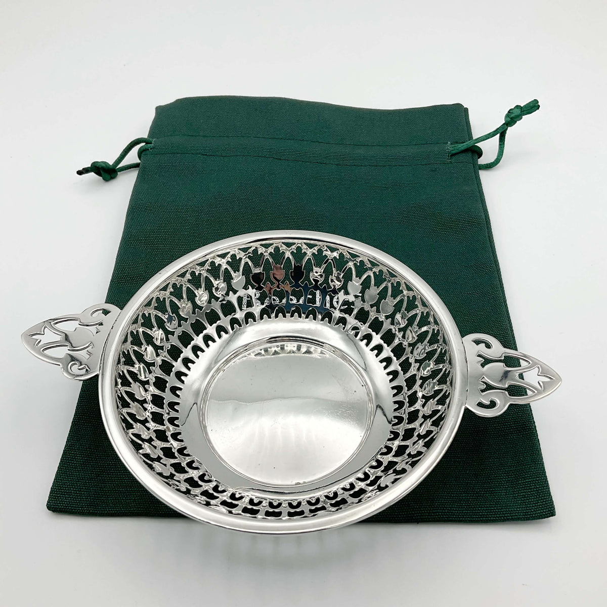 Shiny silver pierced bowl with handles on green cotton gift  bag and a white background
