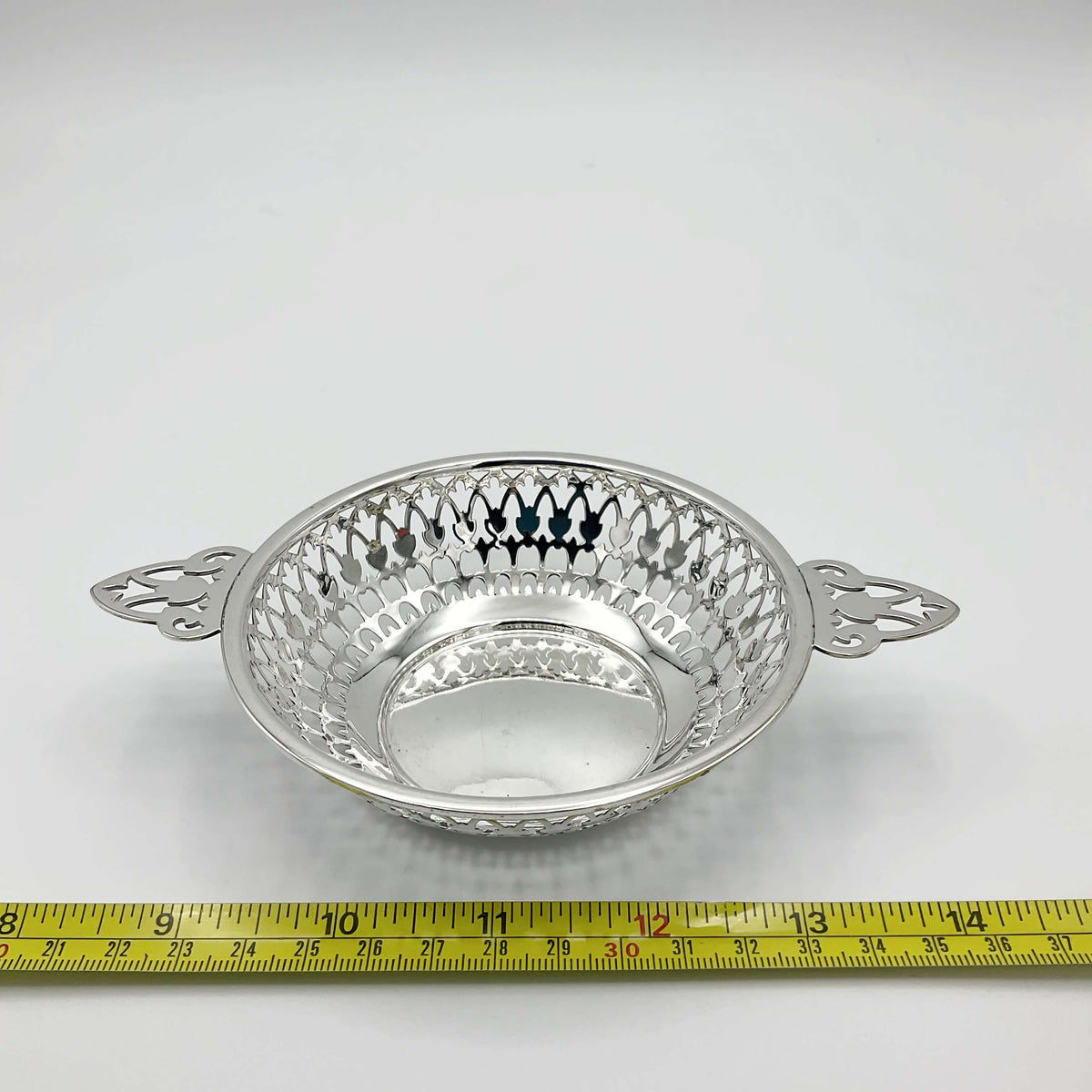 Shiny silver pierced bowl with handles next to tape measure showing width approximately 15cm on a white background