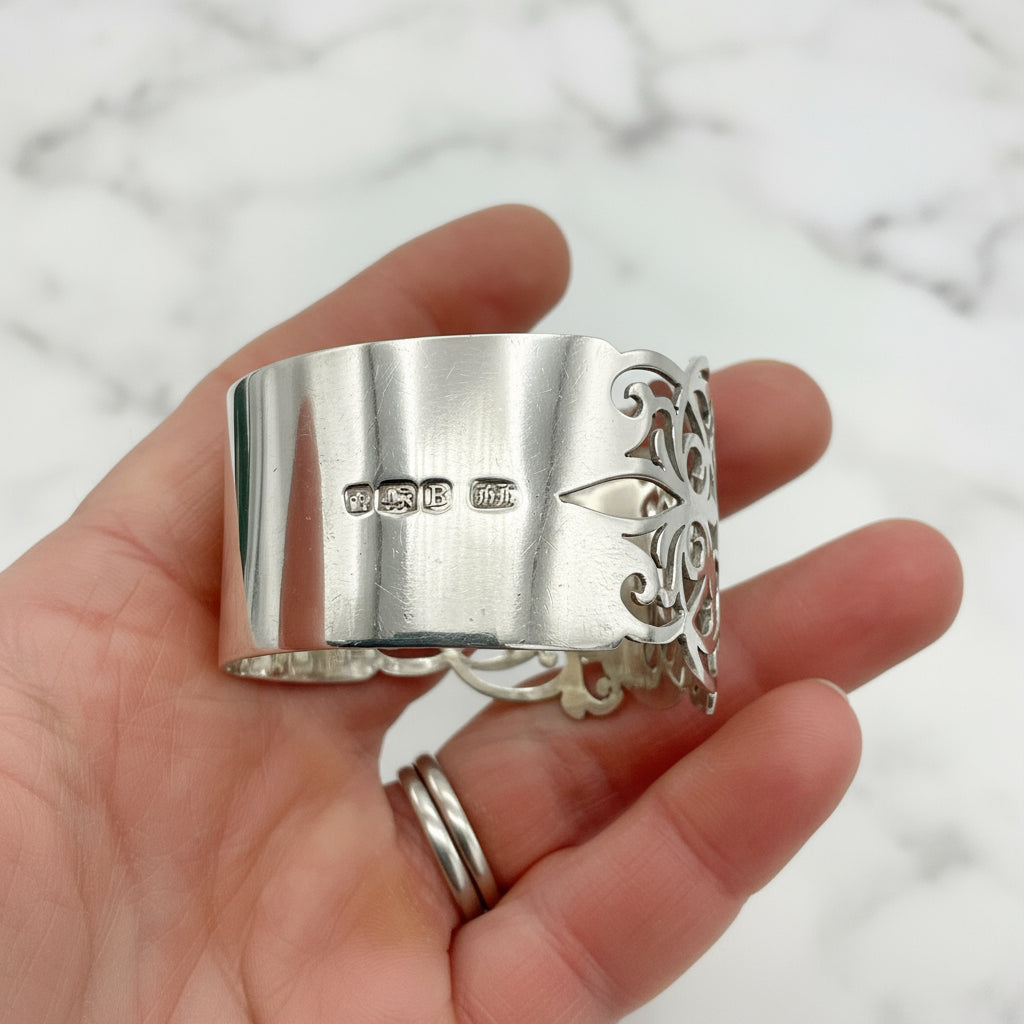 Shiny napkin ring with elaborate sides and hallmarks in a hand on a plain background 