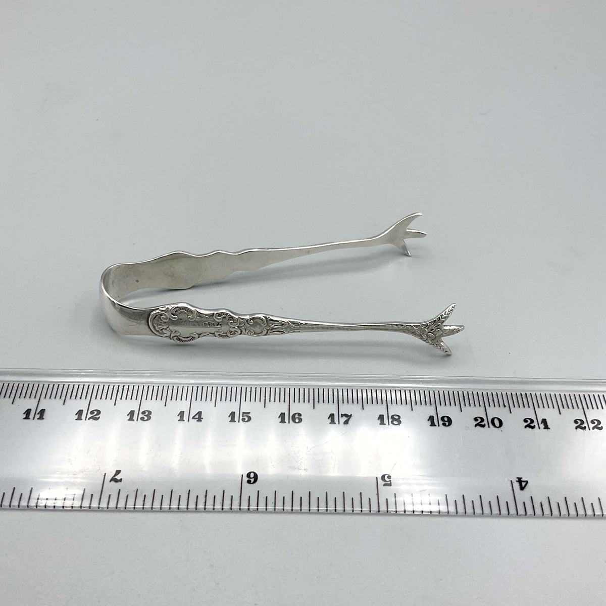 Antique 1902 small sterling silver sugar tongs with ornate design, crafted by Beeches Vintage, resting on a white background.