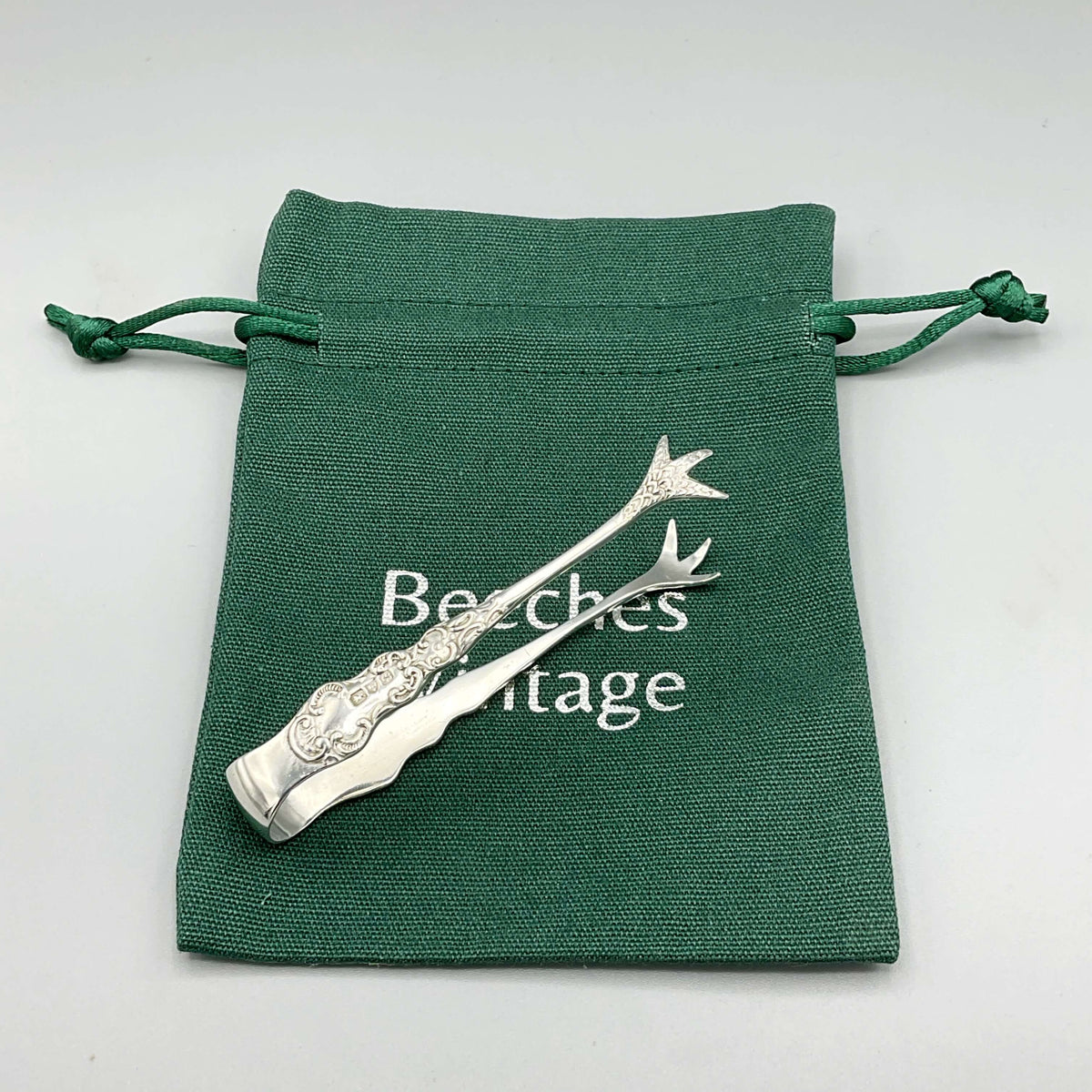 Antique 1902 small sterling silver sugar tongs with ornate design, crafted by Beeches Vintage, on a white background.