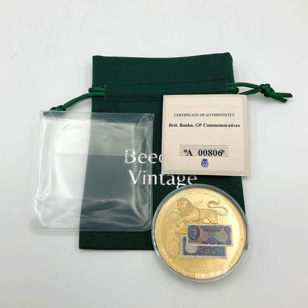 A gold-plated commemorative coin with a banknote sticker on one side, displaying an image of Helmeted Britannia, a lion, and a key. It comes in a green gift bag with a Certificate of Authenticity and is labeled as a collectible.