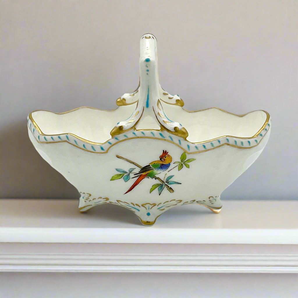 An antique Coalport porcelain china basket with gold and blue trim, featuring a colorful parrot, leaves, and flowers on it. On a white ledge.