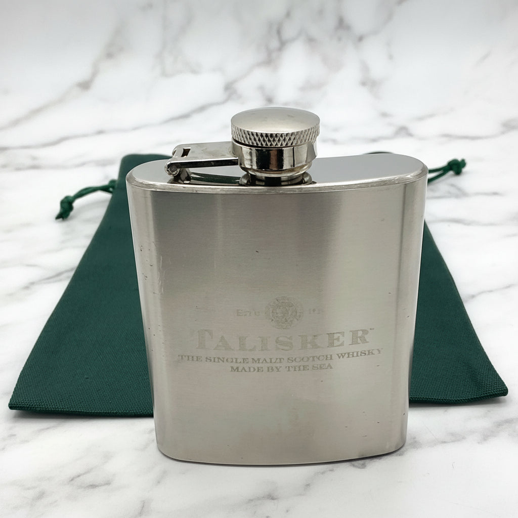 A stainless steel Talisker hip flask with a capacity of 3oz, featuring a brushed design, a hinged screw lid, and the Talisker logo on the front. A green gift bag is also visible in the image.