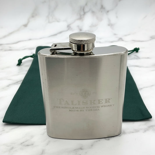A stainless steel Talisker hip flask with a capacity of 3oz, featuring a brushed design, a hinged screw lid, and the Talisker logo on the front. A green gift bag is also visible in the image.