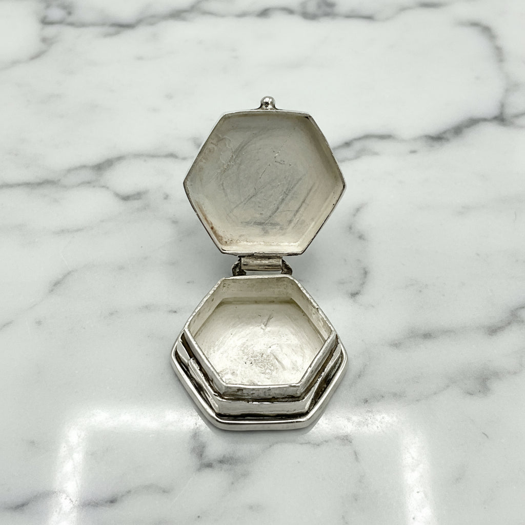 Hexagonal silver pill box with open lid showing the inside on a marble worktop