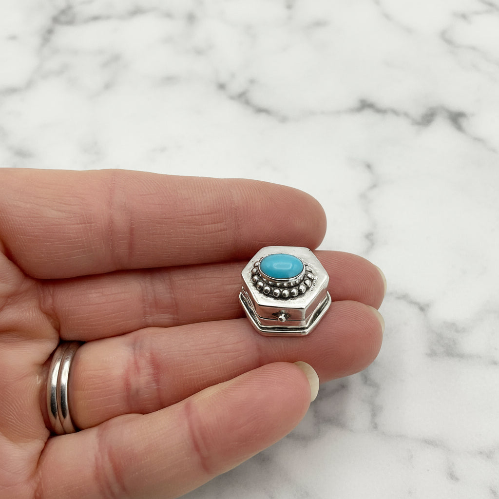 Hexagonal silver pill box with turquoise stone held between fingers on a marble background