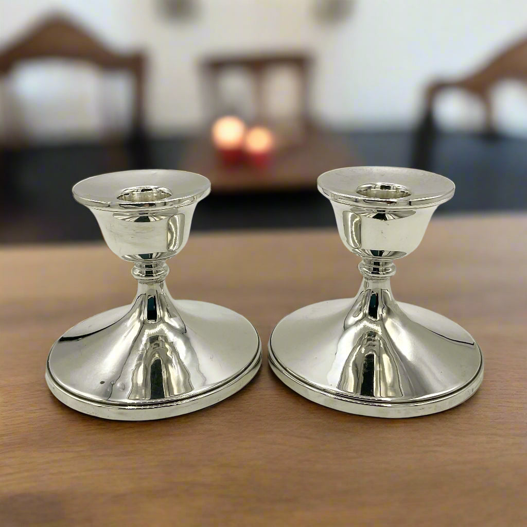 Pair of silver candlestick holders on a wooden table with a table and chairs in the background with lit candles on