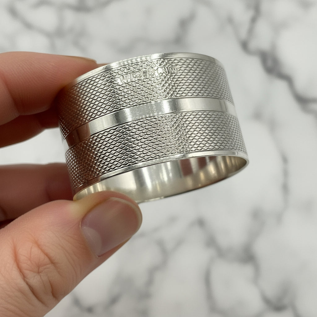The maker’s mark and hallmarks on the side of a silver napkin ring held in fingers with a green gift bag in the background 