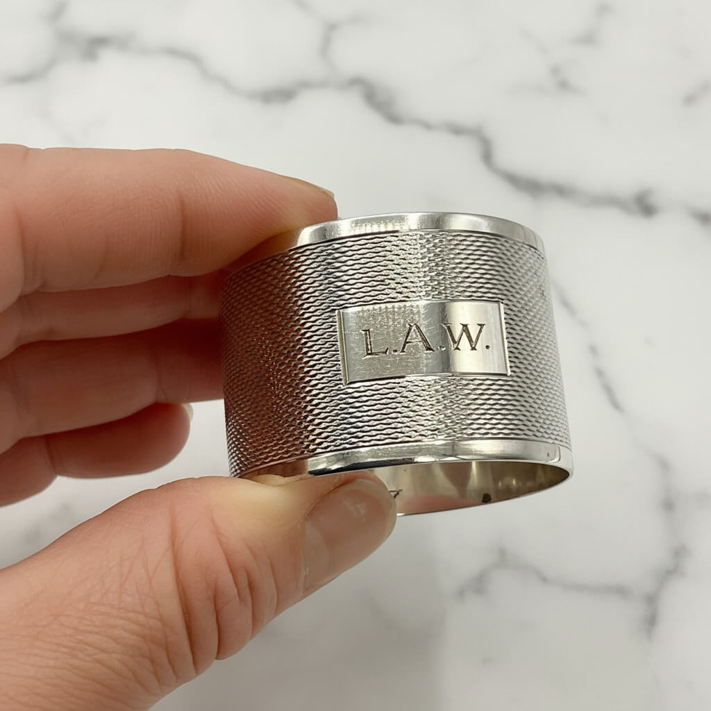 Silver napkin ring with LAW engraved on the front held between fingers with a marble background 