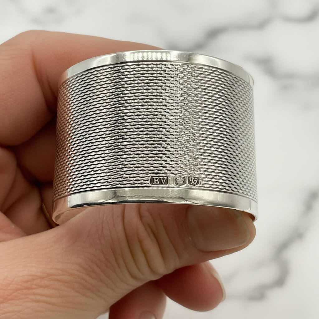 Silver napkin ring with the maker’s mark and hallmarks showing, held between fingers with a white background 
