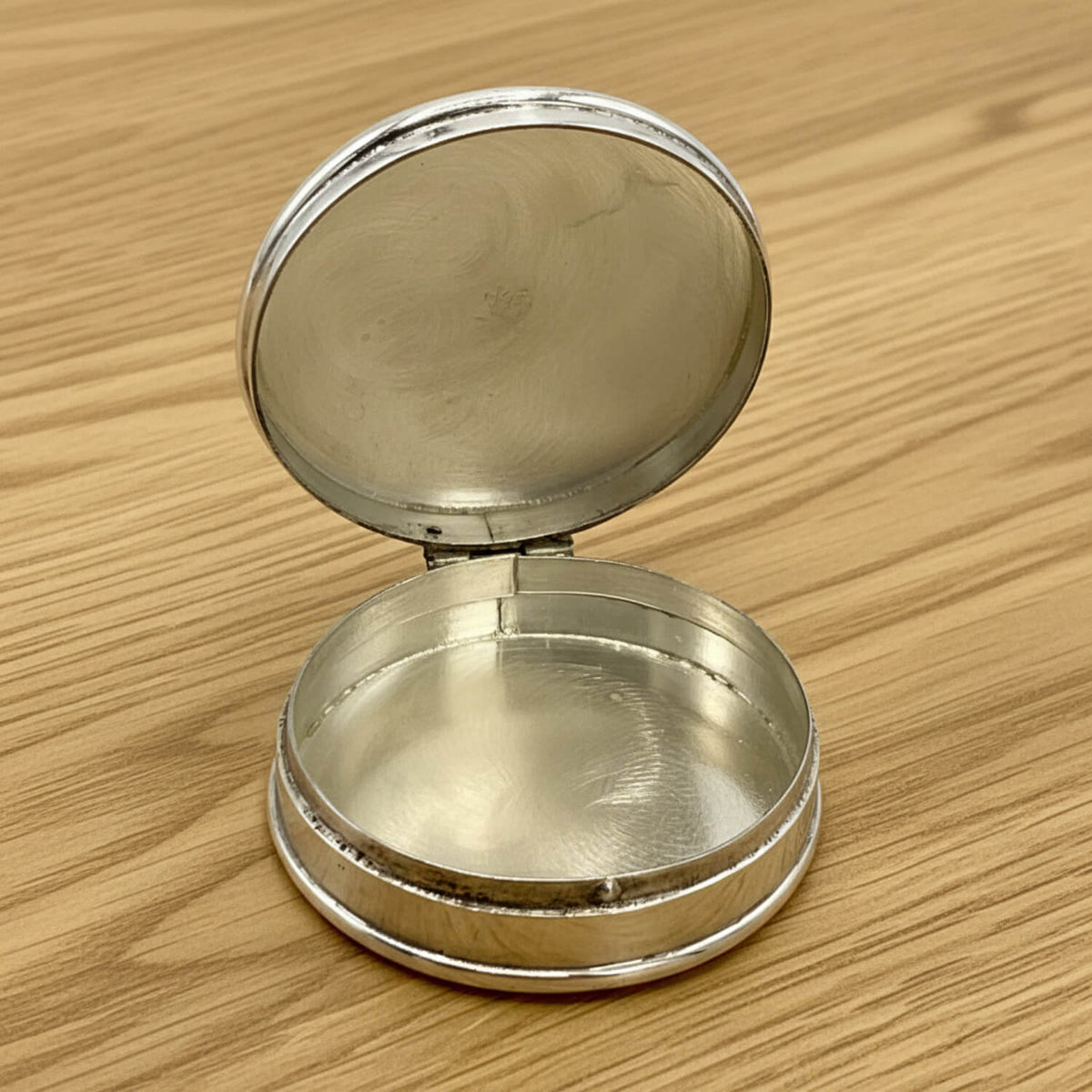 A small round silver  pill box with its lid open, resting on a light brown wooden surface.