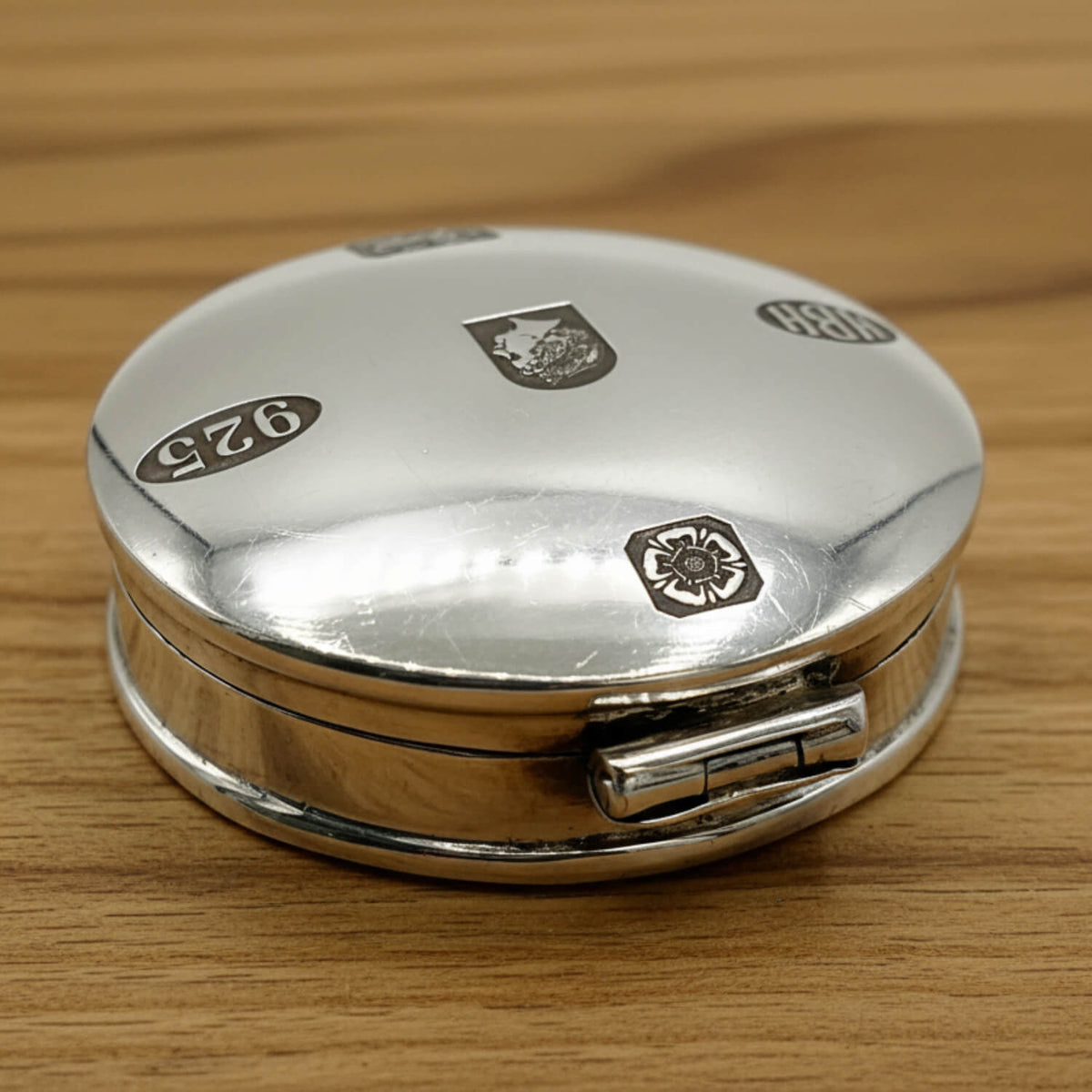 Back of a round sterling silver pill box with engraved hallmarks and a visible hinge, resting on a light brown wooden surface.