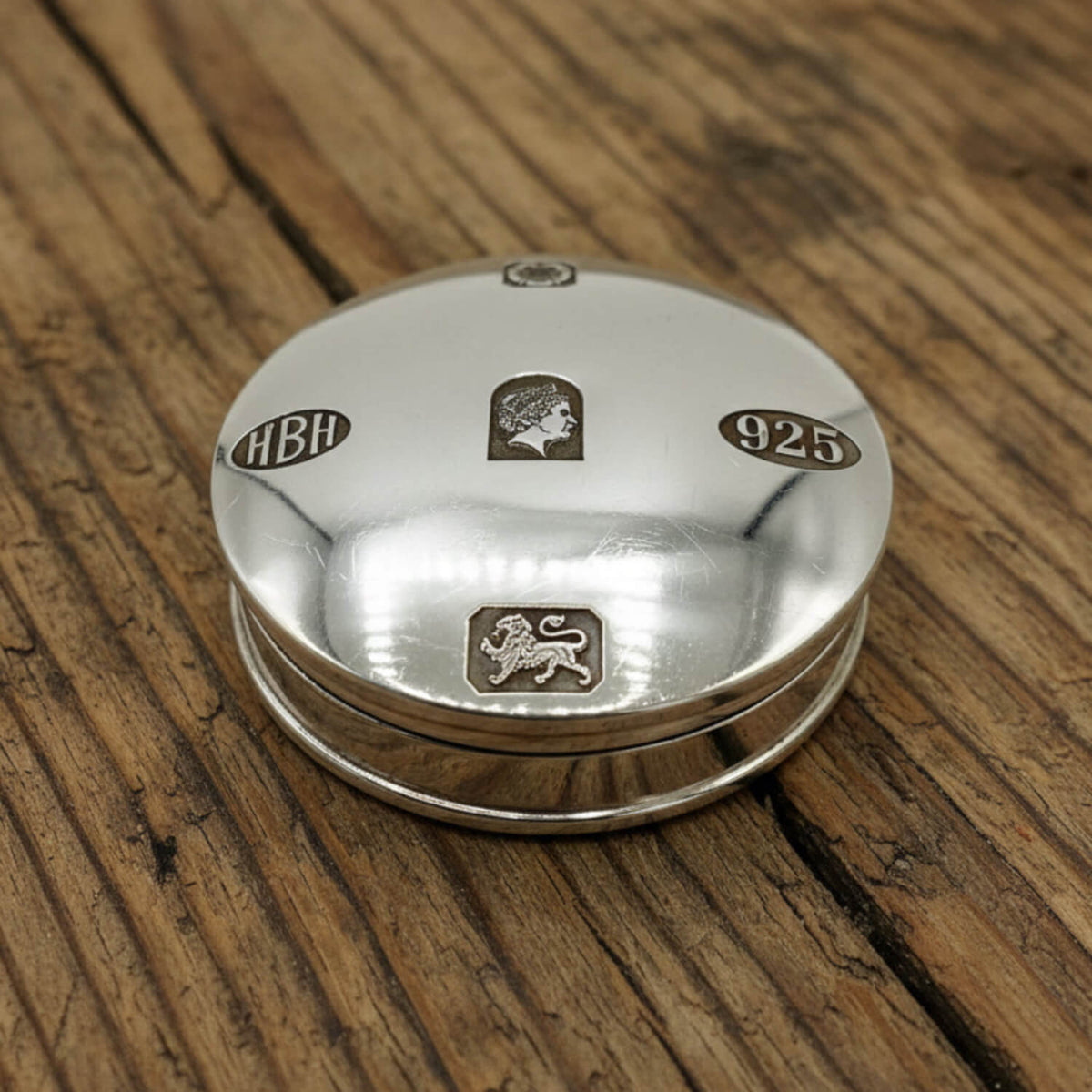 A round sterling silver pill box with engraved details, including the letters 'HBH', a lion passant, and '925', resting on a rustic wooden surface.