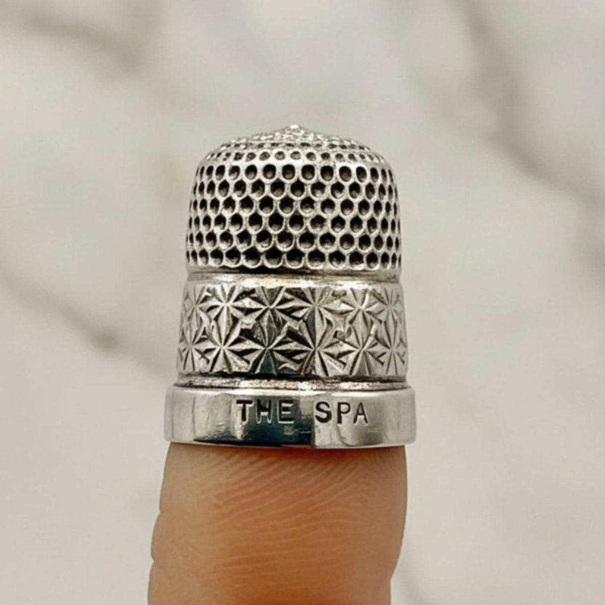 Silver thimble with intricate patterns and 'The Spa' engraving on a finger against a neutral background