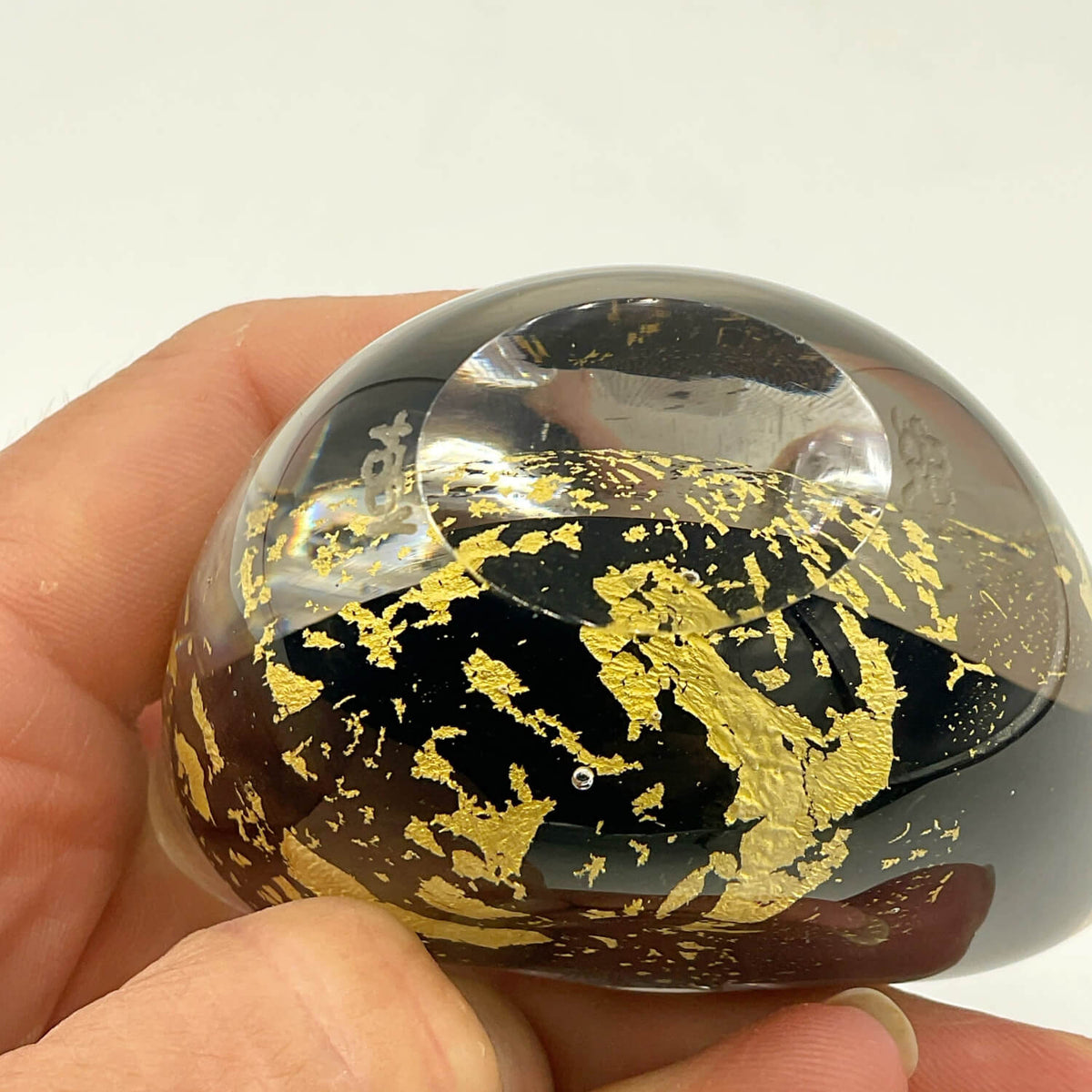 A hand holding a small, oval-shaped clear glass vase filled with black and gold leaf patterns, set against a plain white background.