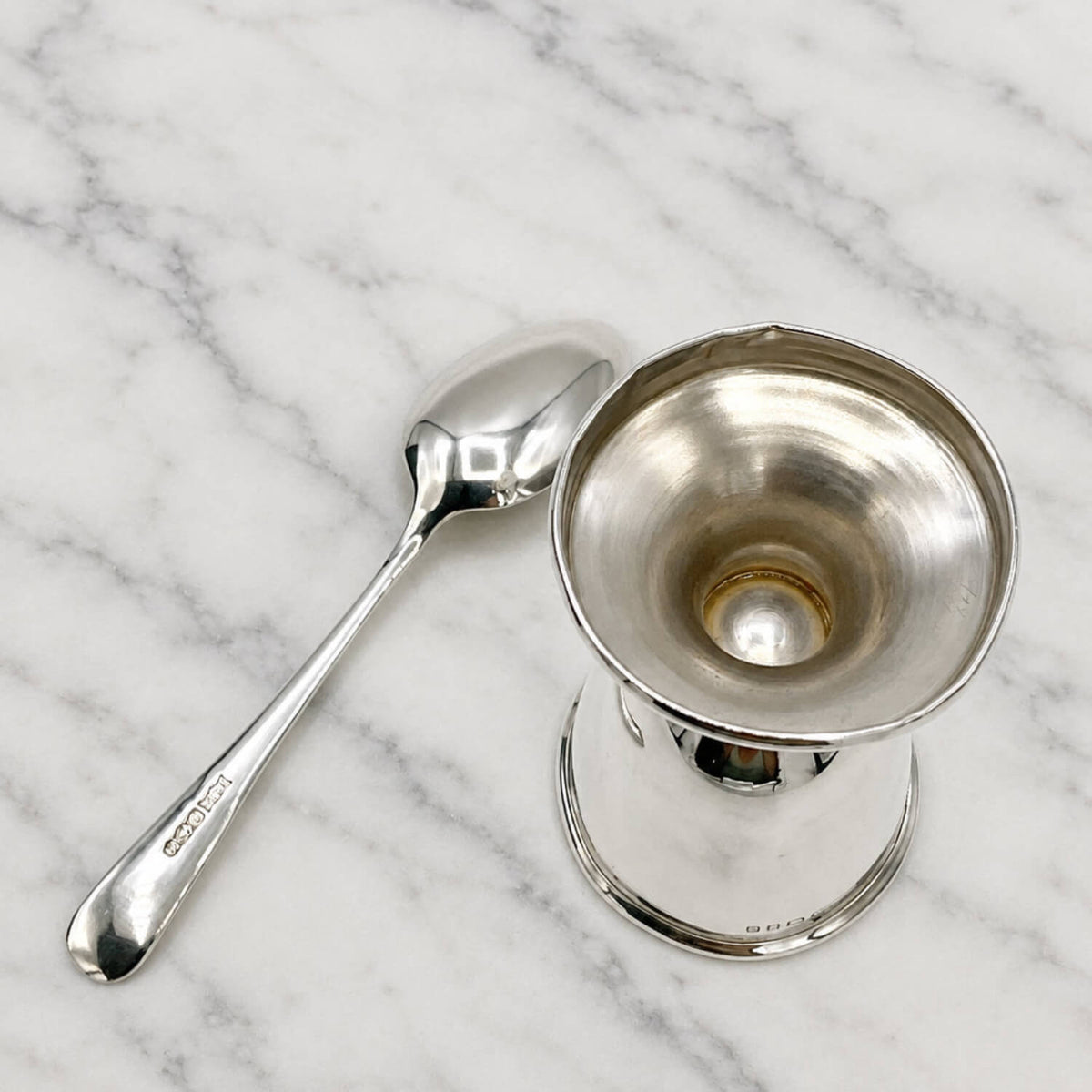 A polished silver egg cup with a small spoon placed beside it on a plain light background.