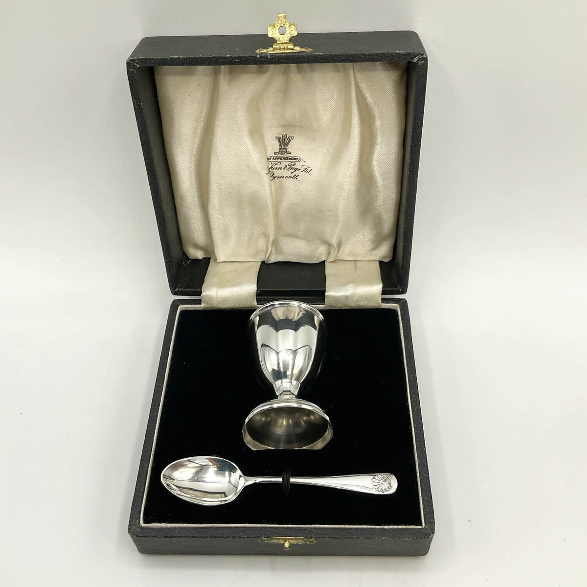 A silver egg cup and spoon presented in an open black gift box with a cream satin lining.