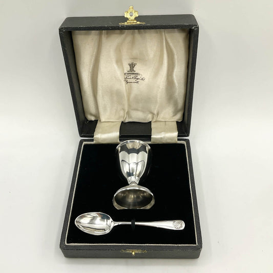 A silver egg cup and spoon presented in an open black gift box with a cream satin lining.
