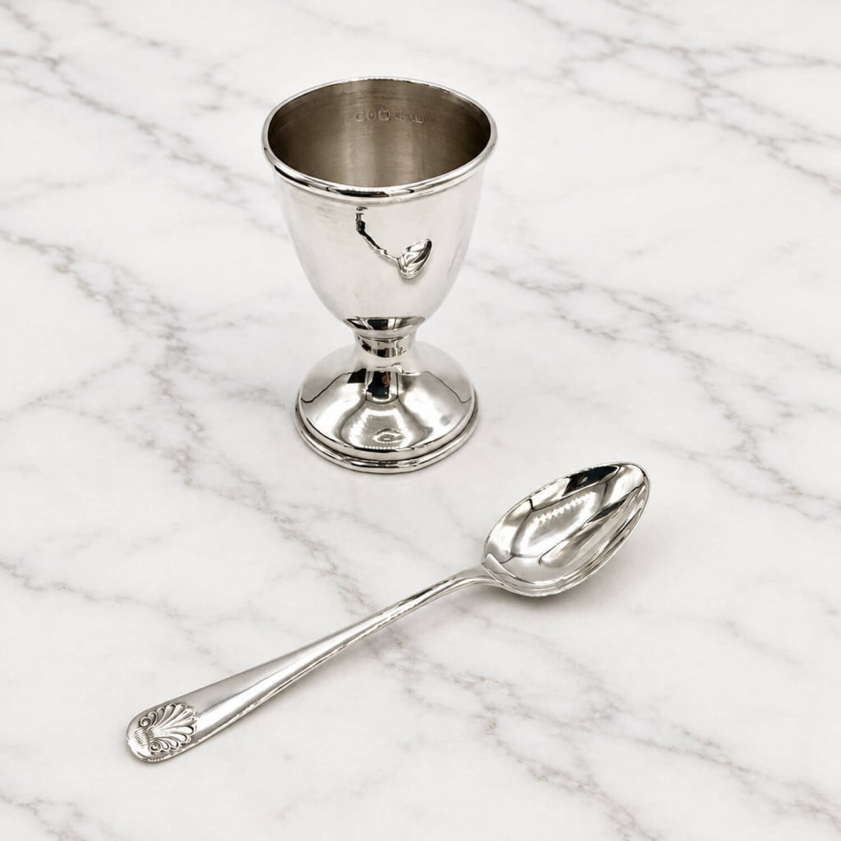 A polished silver pedestal egg cup with a matching silver spoon resting in front of it, set on a white marble surface with gray veining.