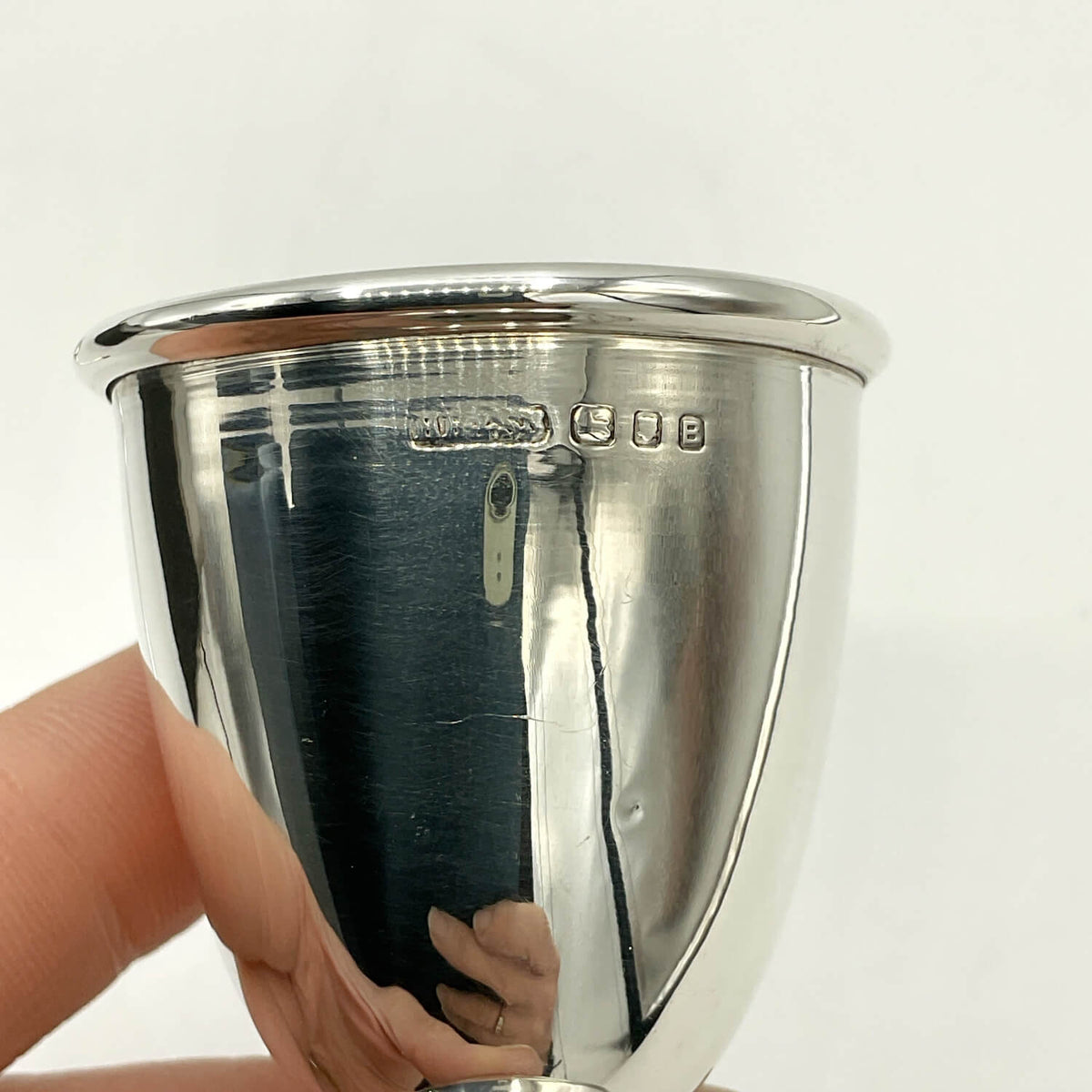 A close-up of a polished silver egg cup being held by a hand, showing a stamped  hallmarks on the side against a plain white background.