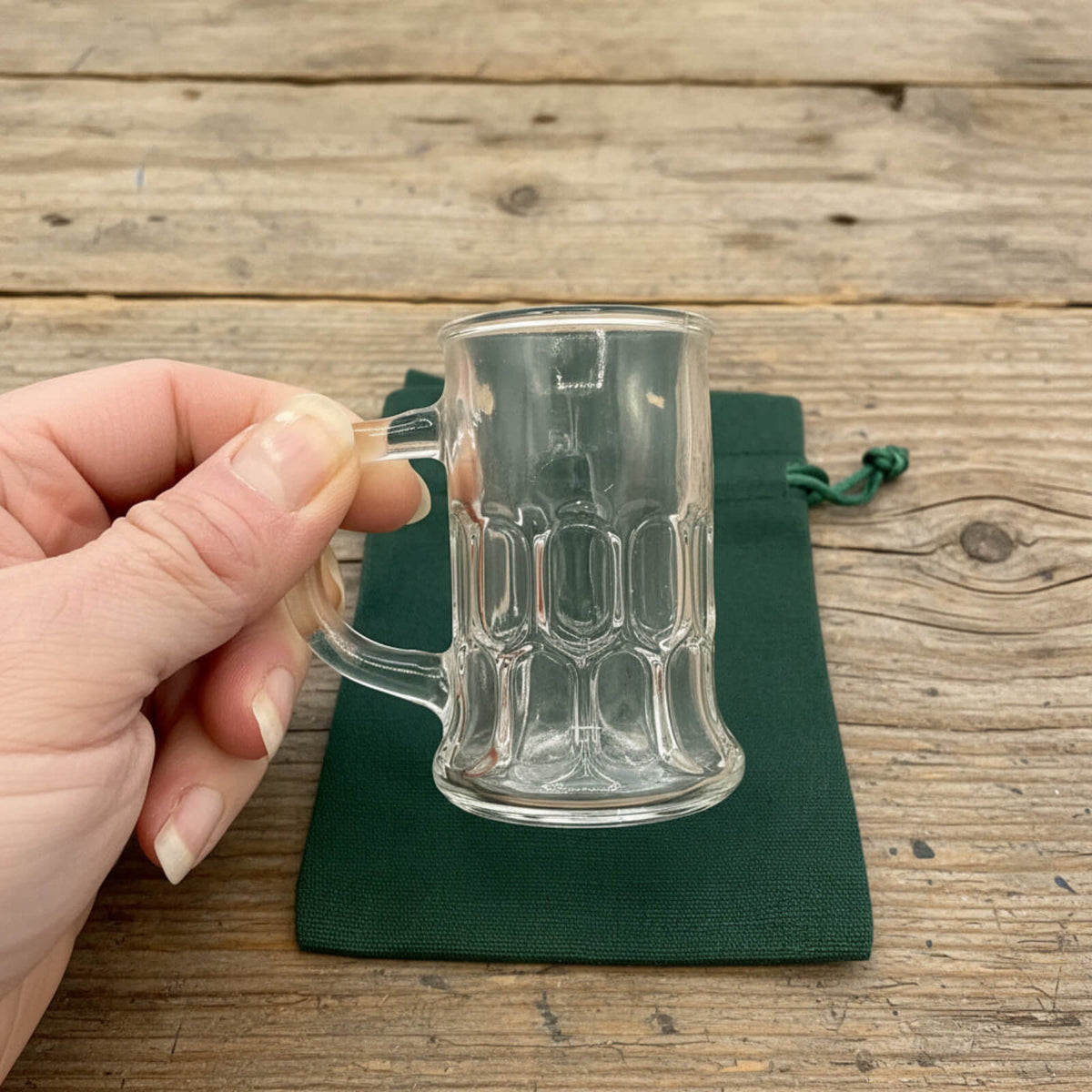 Clear glass mug held by a hand on a wooden surface with a green drawstring bag.