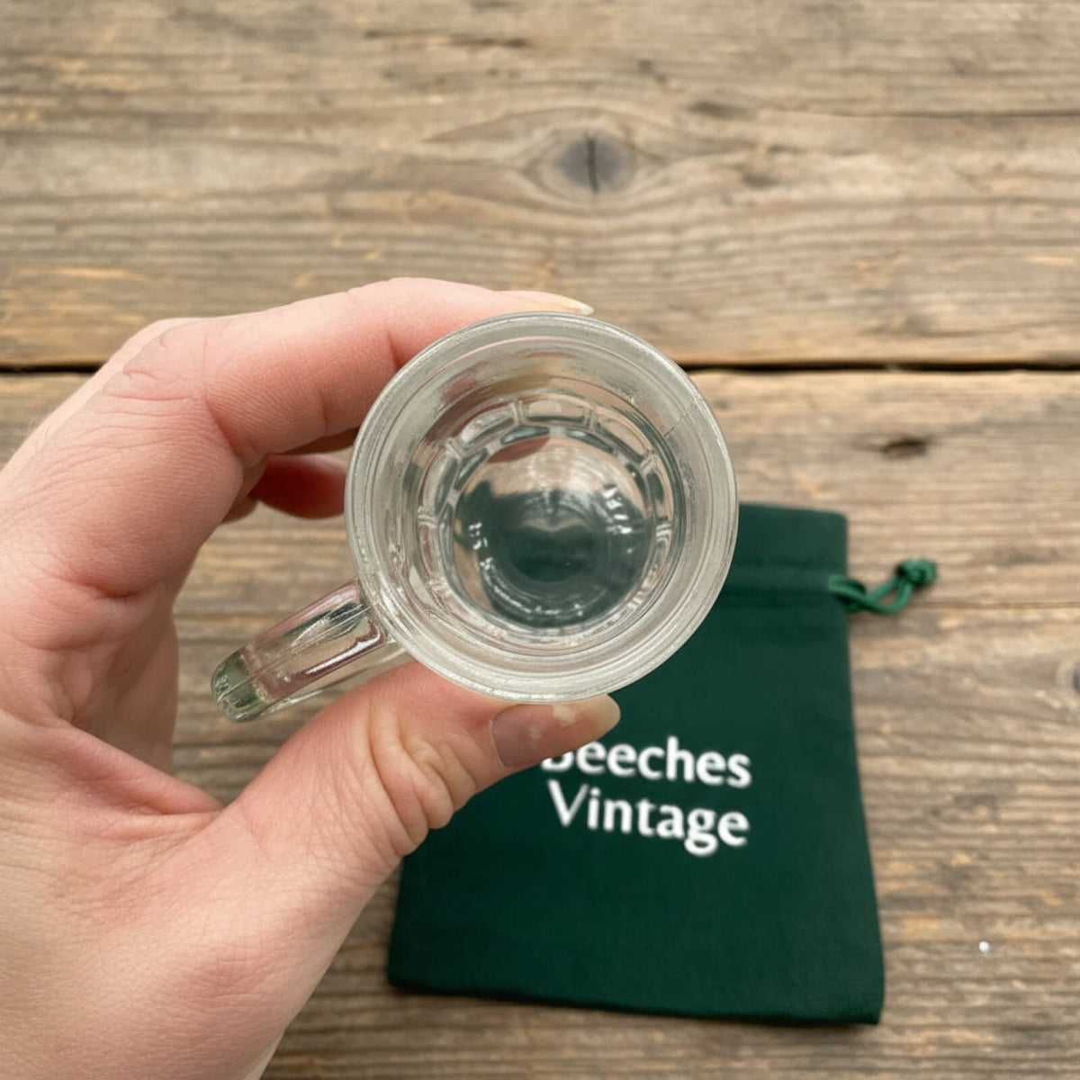 Clear glass mug held by a hand on a wooden surface with a green pouch labeled 'Beeches Vintage' in the background.