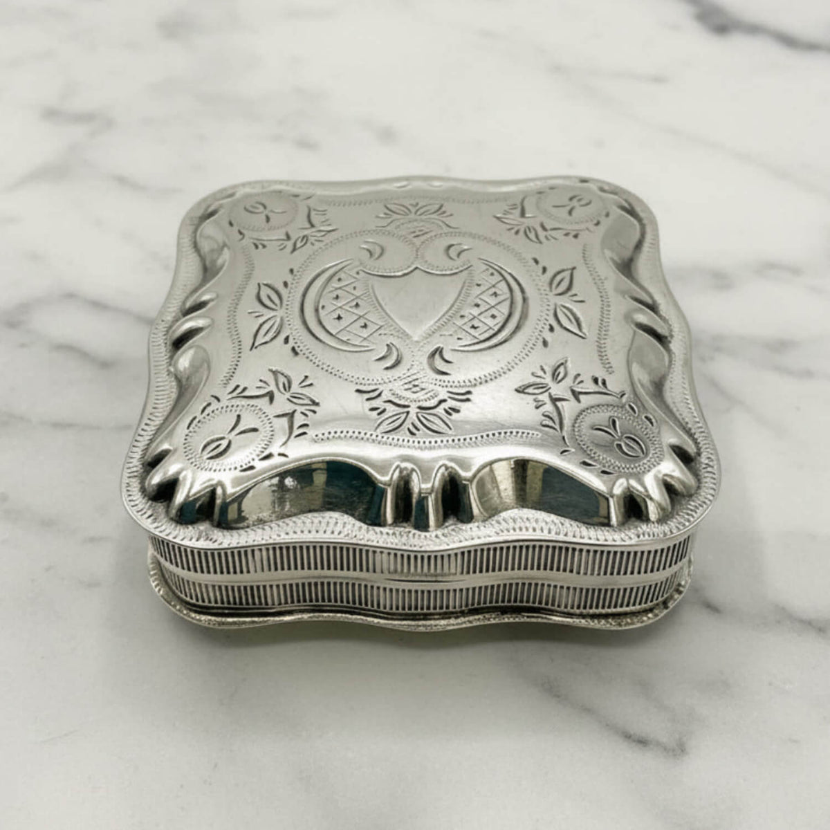 An ornate sterling silver jewelry box with a decorative lid featuring floral engravings and a central crest, resting on a white marble surface.
