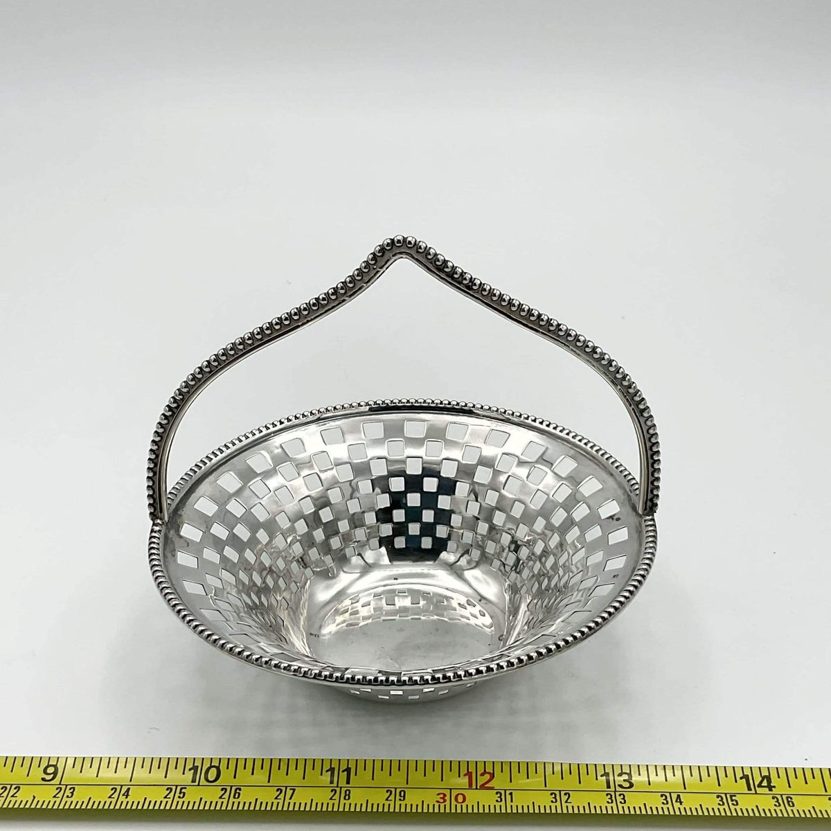 Silver basket with a handle on a white background, measured against a tape measure.