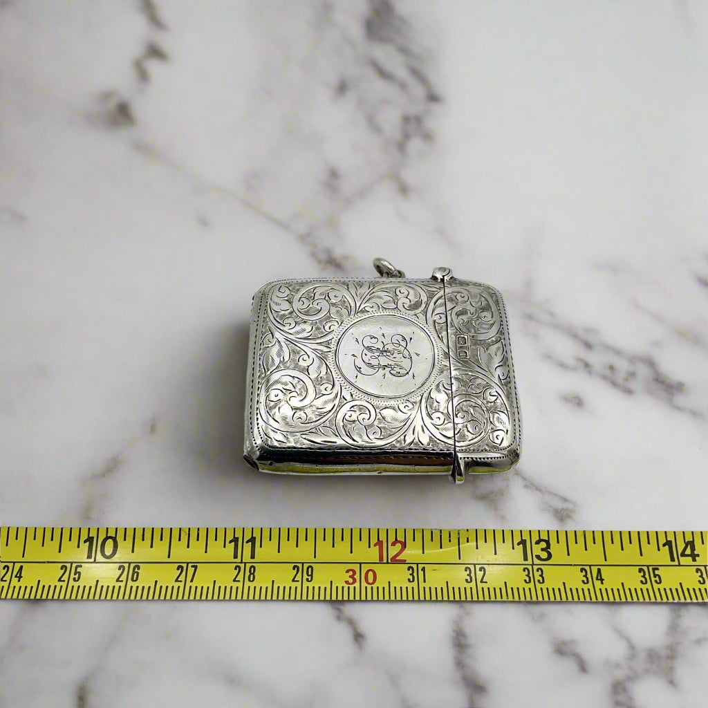 Silver Vesta case with intricate patterns on a marble surface, measured by a yellow tape measure 