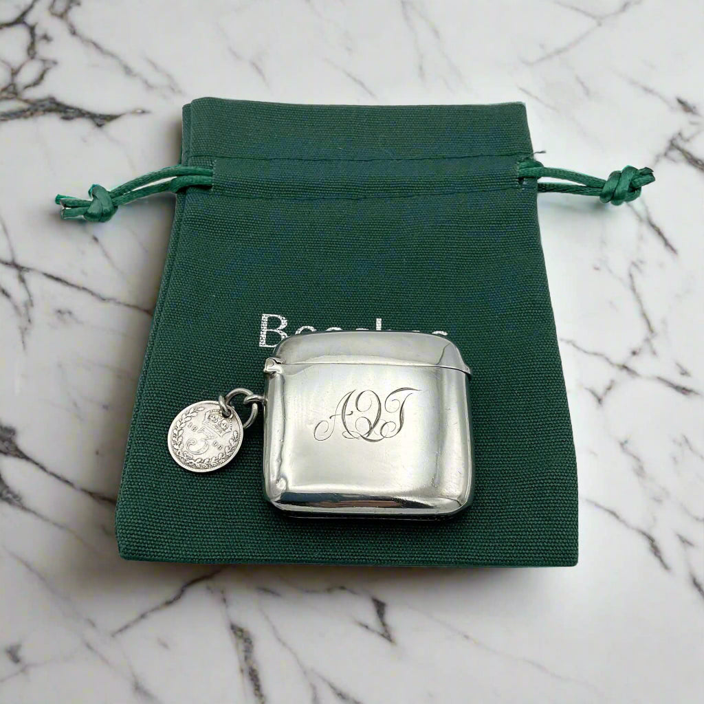 Silver vesta with monogram and round coin on a green drawstring bag with 'Beeches Vintage’ branding, on a marble surface.
