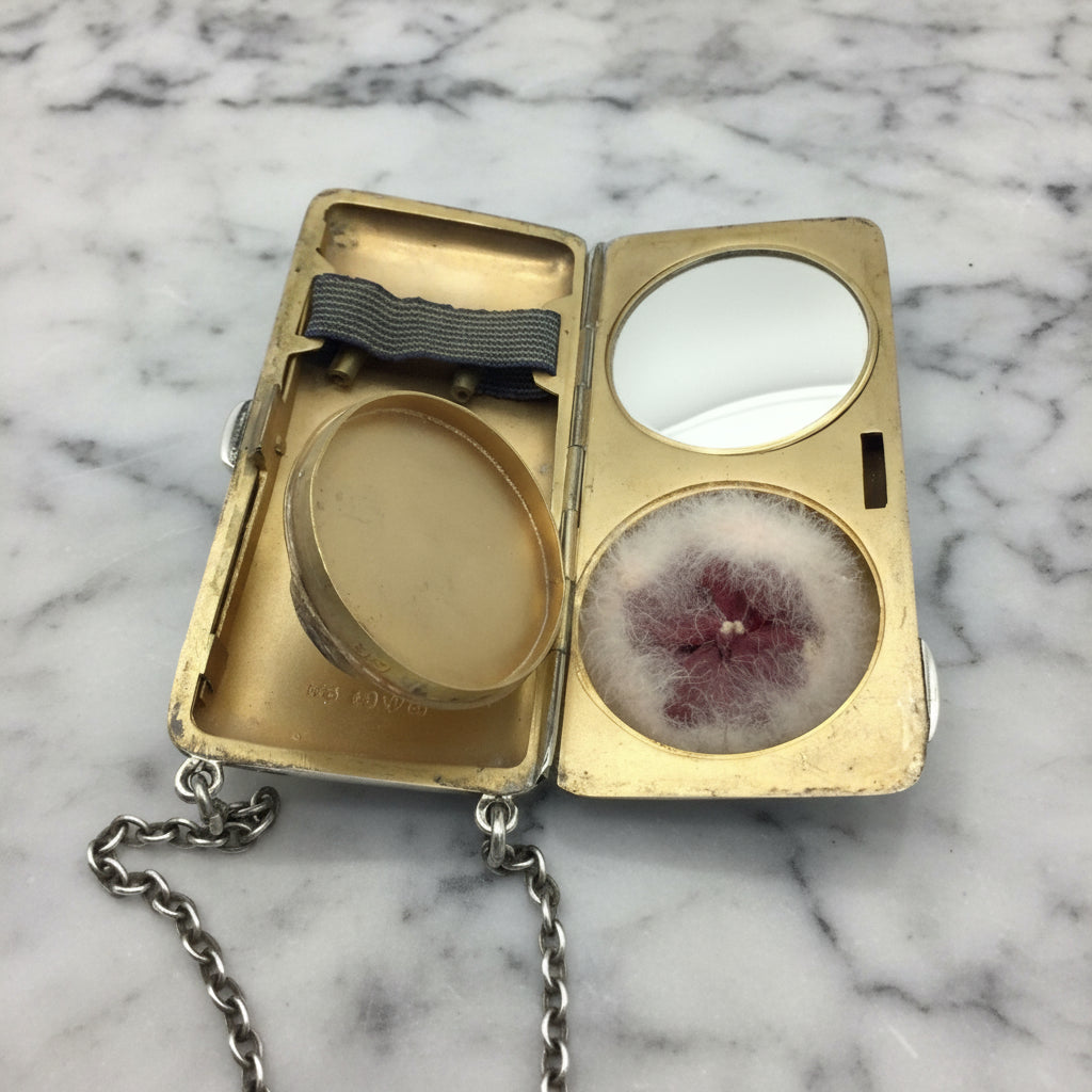 gilded golden inside of case showing a mirror, powder area with an open lid, a powder puff and chain on a marble surface