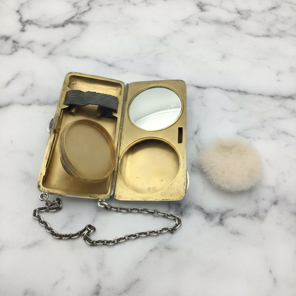 gilded golden inside of case showing a mirror, powder area and chain, with the unused powder puff at the side on a marble surface