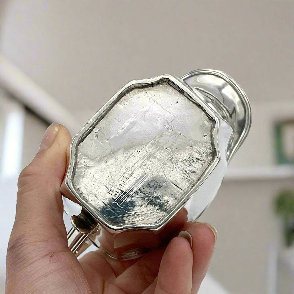 Base of antique silver jug held in a hand with a room background 