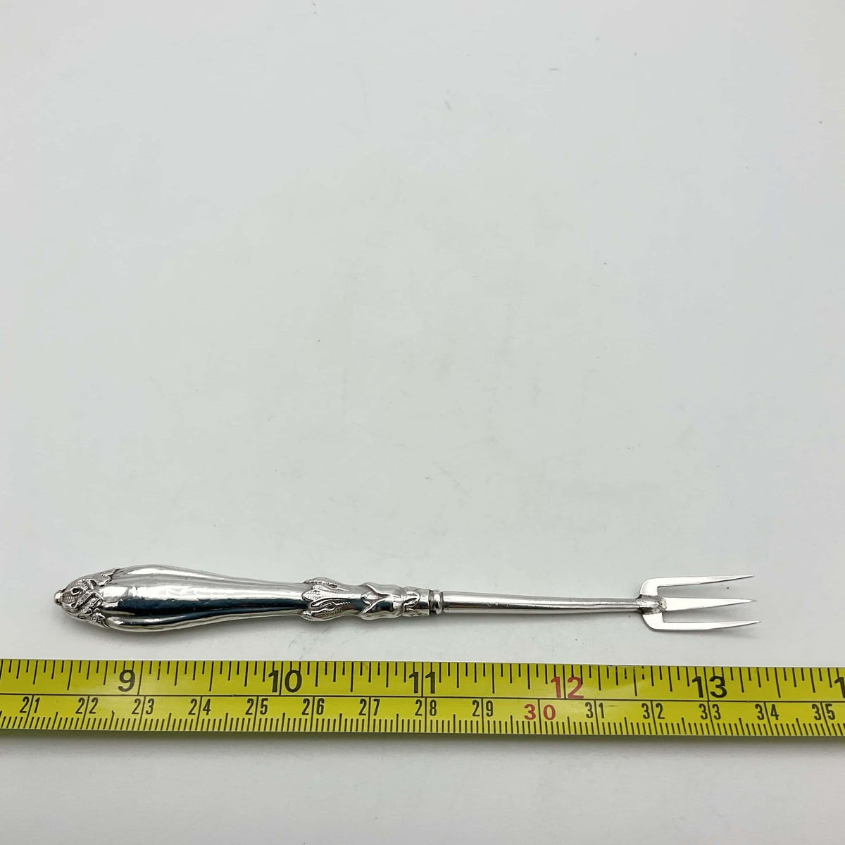 Antique silver plated pickle fork with an ornate handle next to a tape measure