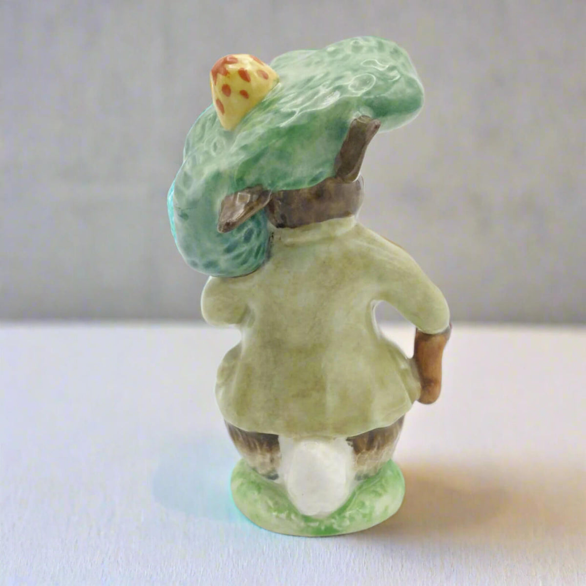 Small ceramic figure of a person wearing a green hat and holding a shoe on a white surface.