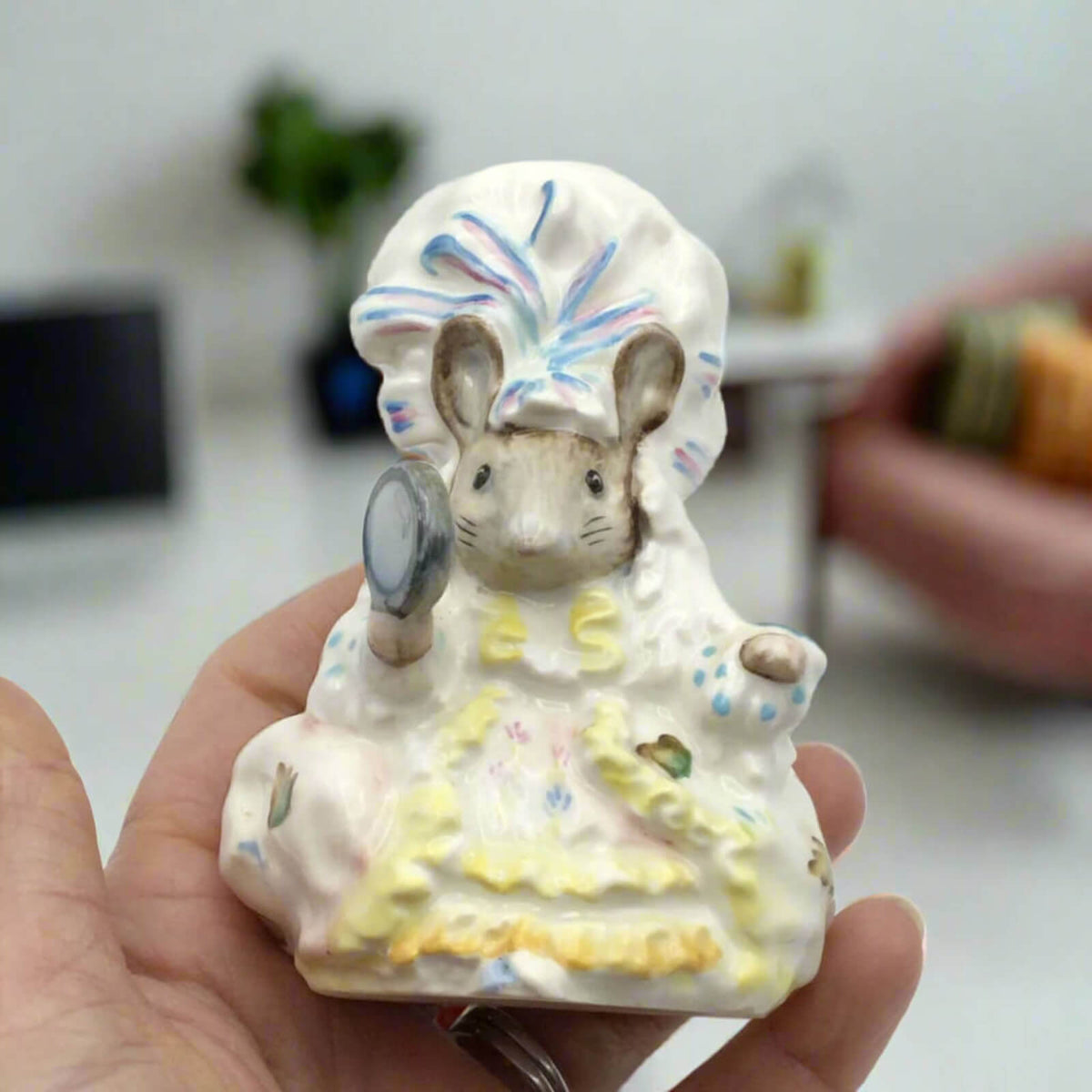 Small ceramic figurine of a mouse with a colorful hat held in a hand.