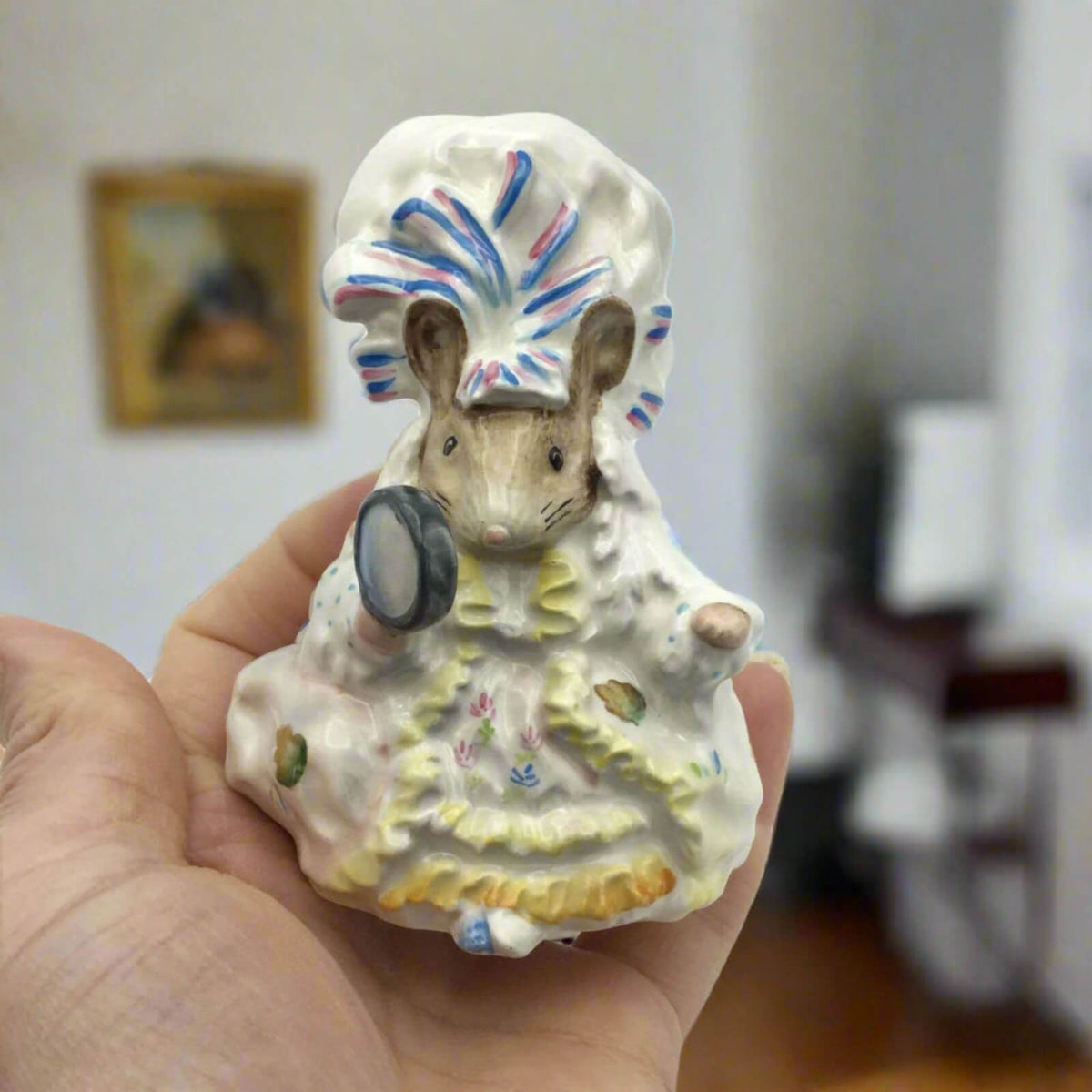 Small ceramic figure of a mouse in a headdress held in a hand with a blurred indoor background.