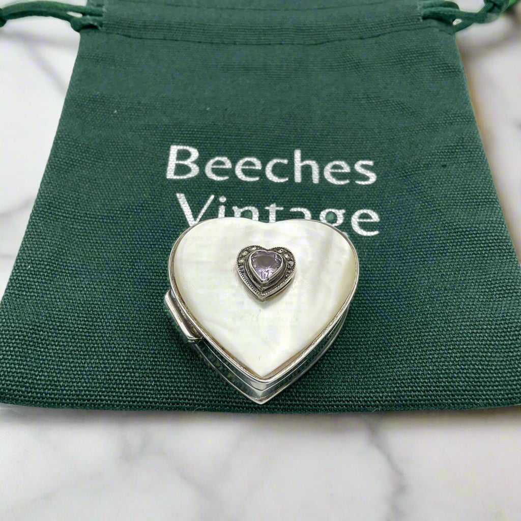 Heart shaped silver pill box with an amethyst and mother of pearl lid on a green bag on a marble background.