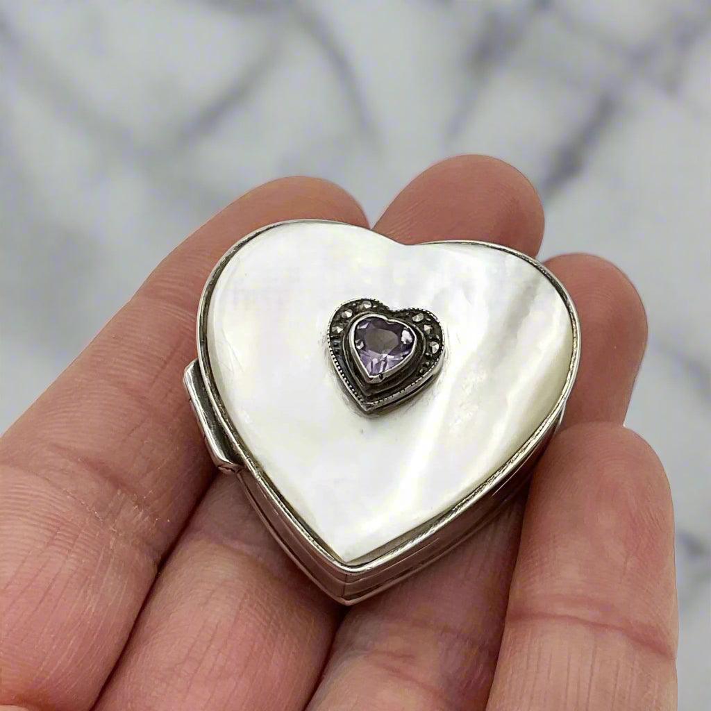 Heart shaped silver pill box with an amethyst and mother of pearl lid in a hand on a marble background.