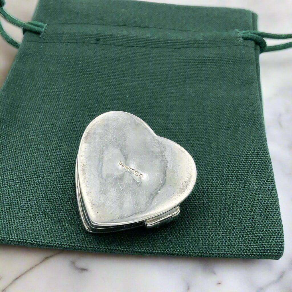 Base of Heart shaped silver pill box on a green bag on a marble background.