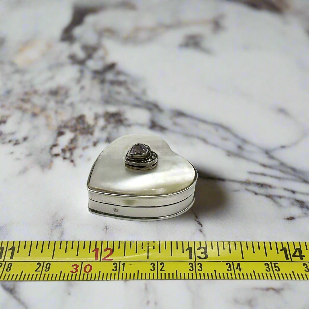 Heart shaped silver pill box with an amethyst and mother of pearl lid next to a tape measure showing the length as approx 3cm on a marble background.