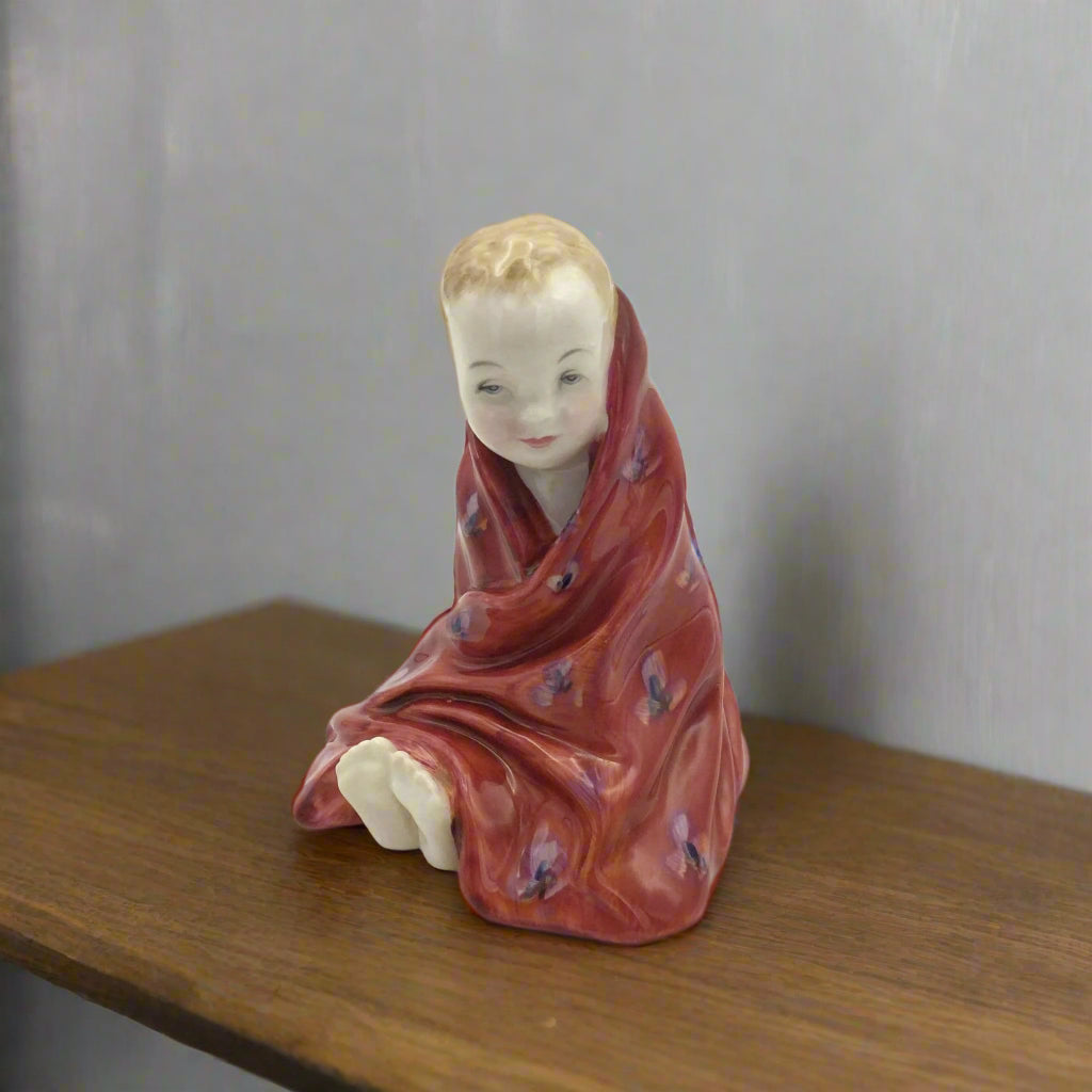 Vintage ceramic figurine of a child wrapped in a red cloth on a wooden shelf.