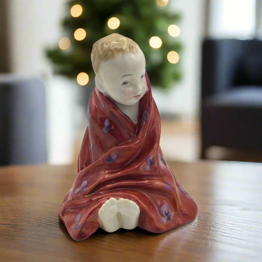 Ceramic figurine of a child wrapped in a red cloth on a wooden surface with a Christmas tree in the background