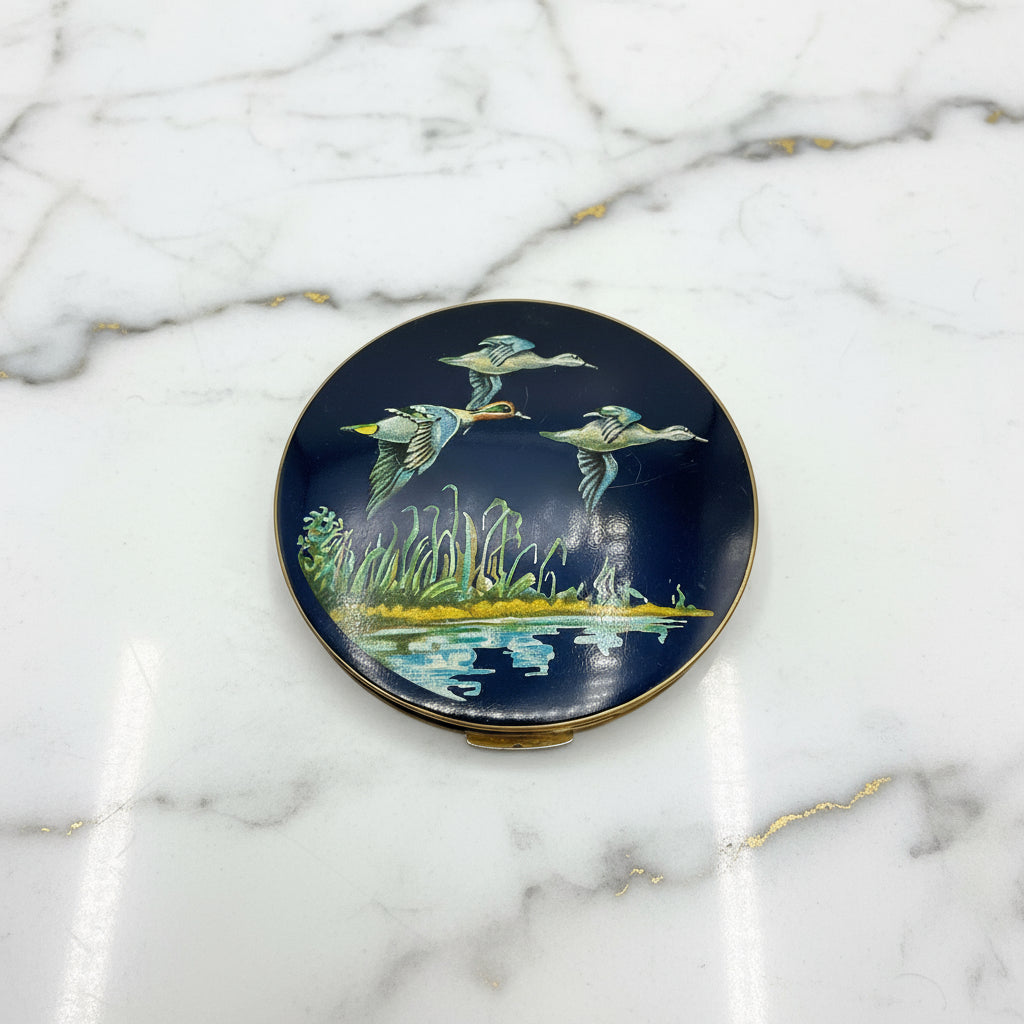 beautiful enamel powder compactfeaturing three ducks flying over water on a mirror compact lid on a marble surface
